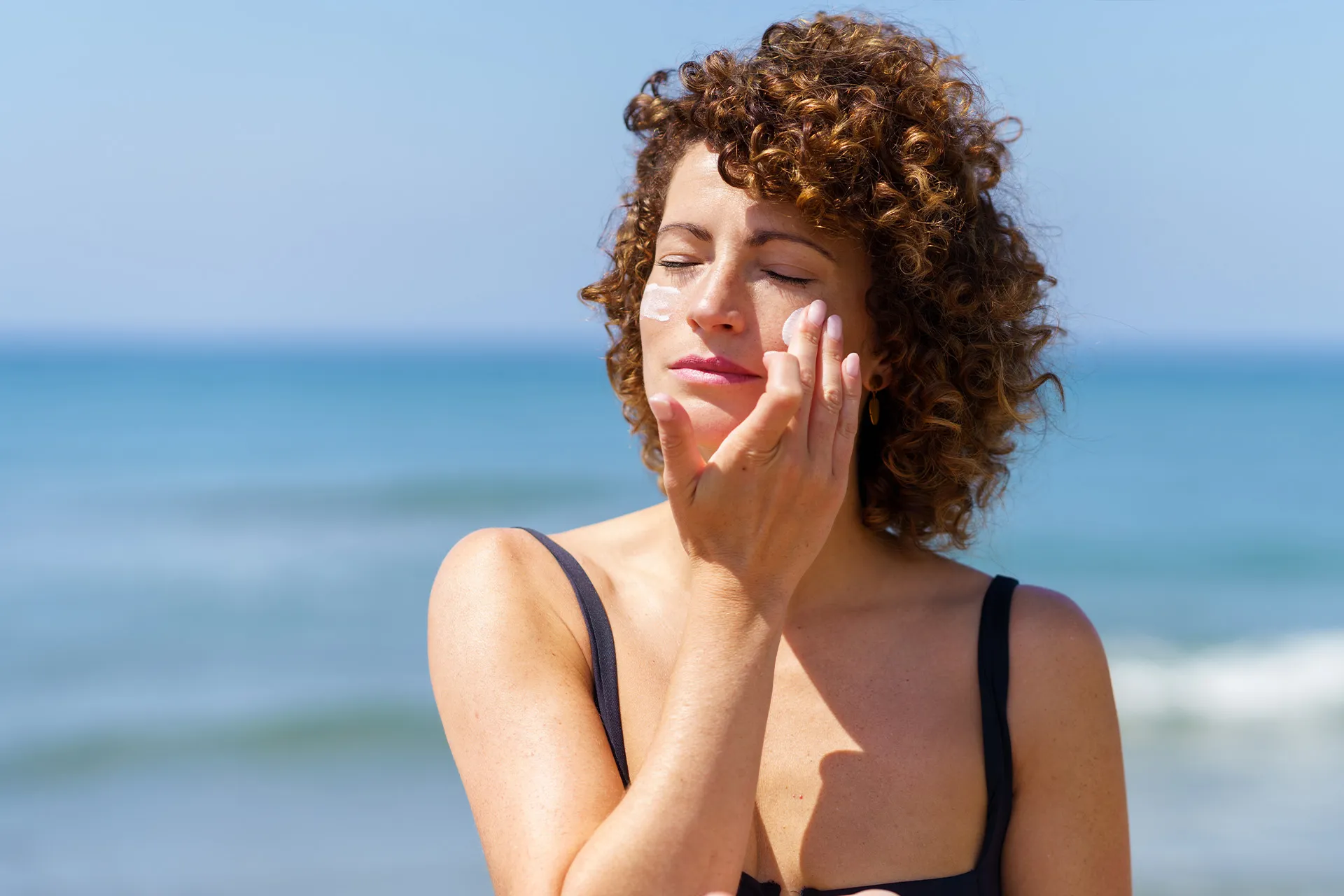 Frau mit lockigen Haaren trägt am Strand Sonnenschutzcreme als letzten Schritt ihrer Pflegeroutine auf